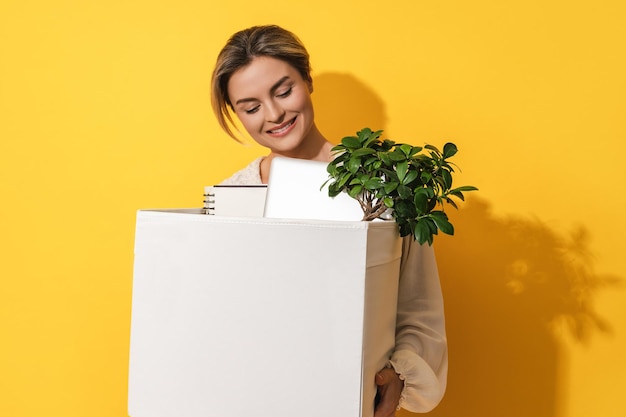 Mujer feliz sosteniendo una caja con artículos personales después de la promoción laboral contra el fondo amarillo