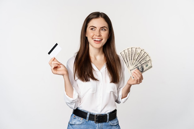 Mujer feliz sosteniendo dinero y tarjeta de crédito con cara pensativa, concepto de préstamo y microcrédito, de pie sobre fondo blanco.