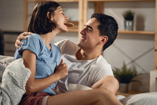 Mujer feliz sosteniendo una galleta en la boca y compartiéndola con su novio en el dormitorio