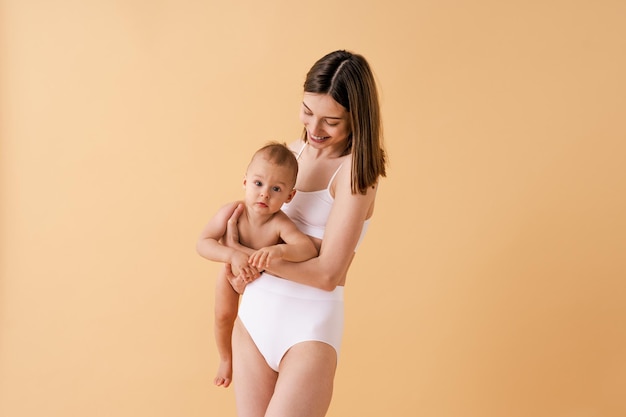 Mujer feliz sosteniendo a su bebé con fondo de color - Mujer joven usando ropa interior cuidando a su hijo pequeño - Embarazo, maternidad, personas y concepto de expectativa