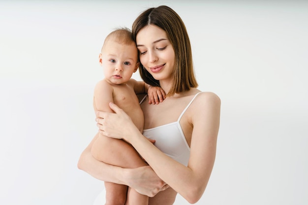 Mujer feliz sosteniendo a su bebé con fondo de color - Mujer joven usando ropa interior cuidando a su hijo pequeño - Embarazo, maternidad, personas y concepto de expectativa