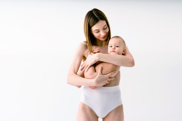 Mujer feliz sosteniendo a su bebé con fondo de color - Mujer joven usando ropa interior cuidando a su hijo pequeño - Embarazo, maternidad, personas y concepto de expectativa