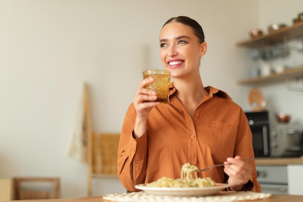 Foto mujer feliz sosteniendo un vaso de agua comiendo espaguetis caseros sentado a la mesa en la cocina y mirando