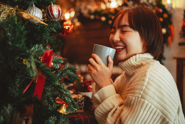 La mujer feliz sostiene una taza de café y huele aromático cerca de la decoración del árbol de Navidad.
