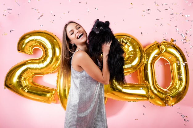 Mujer feliz con spitz japonés perro negro y confeti y globos de oro 2020 año nuevo aislados en rosa