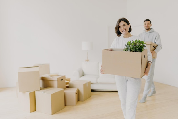 La mujer feliz y su esposo llevan cajas con pertenencias personales ocupadas durante la reubicación en otro lugar para vivir, ingresan a un nuevo hogar, se mudan juntos en una casa grande y desempacan las cosas del hogar