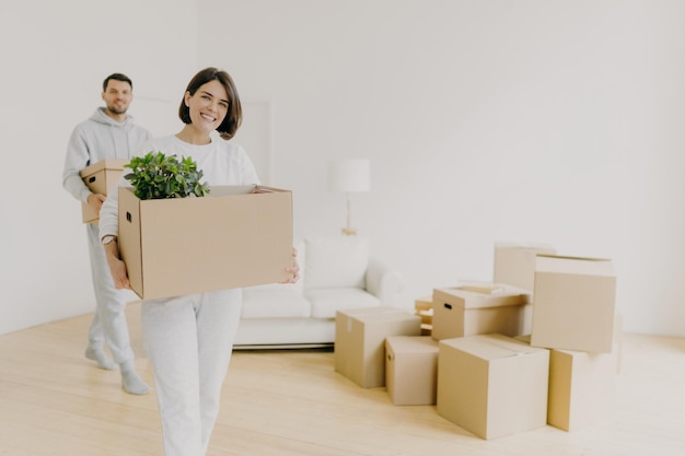 La mujer feliz y su esposo llevan cajas con pertenencias personales ocupadas durante la reubicación en otro lugar para vivir, ingresan a un nuevo hogar, se mudan juntos en una casa grande y desempacan las cosas del hogar