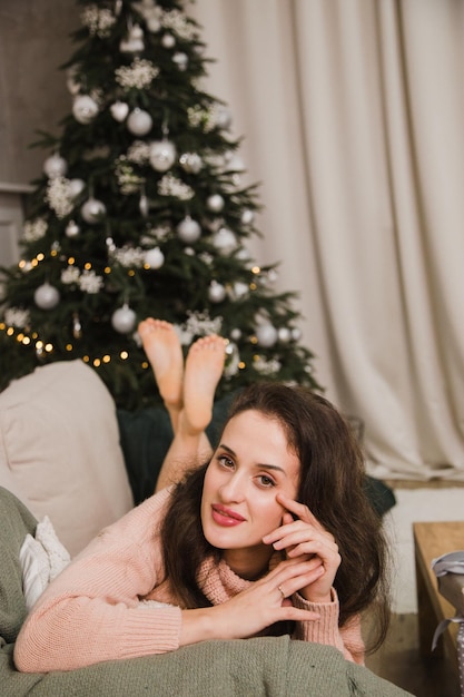 Mujer feliz en un suéter se encuentra en el sofá con el telón de fondo de un árbol de Navidad Año Nuevo