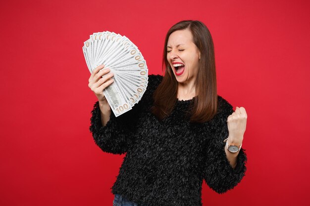 Mujer feliz en suéter de piel gritando, haciendo gesto de ganador mantenga ventilador de dinero en billetes de dólar, dinero en efectivo aislado sobre fondo rojo. Personas sinceras emociones, concepto de estilo de vida. Simulacros de espacio de copia.