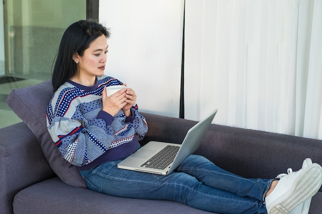 Mujer feliz con la taza de café caliente que pone en el sofá cómodo que usa la computadora portátil