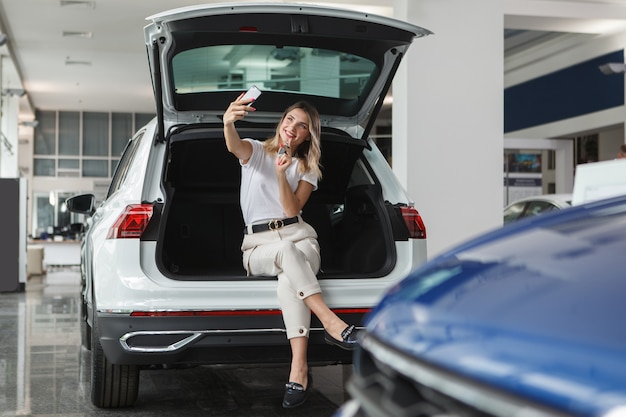 Mujer feliz tomando autorretratos con la llave del coche de su nuevo auto, sentado en el maletero del SUV