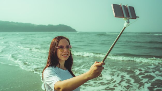 Mujer feliz tomando selfie cerca del mar Desde arriba mujer alegre usando monopie con teléfono inteligente para tomar selfie cerca del mar ondulante en la playa