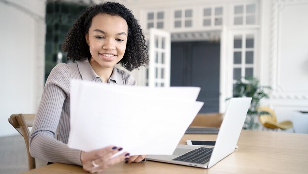 Una mujer feliz trabaja con documentos, una secretaria se sienta en un espacio de coworking de oficina y realiza tareas en una computadora portátil