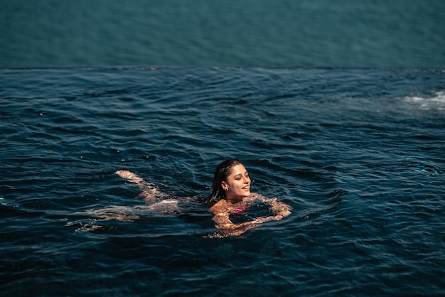 Mujer feliz en traje de baño nadando en la piscina infinita frente al mar