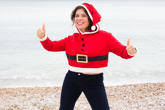 Mujer feliz en traje de santa claus en la playa mostrando los pulgares para arriba