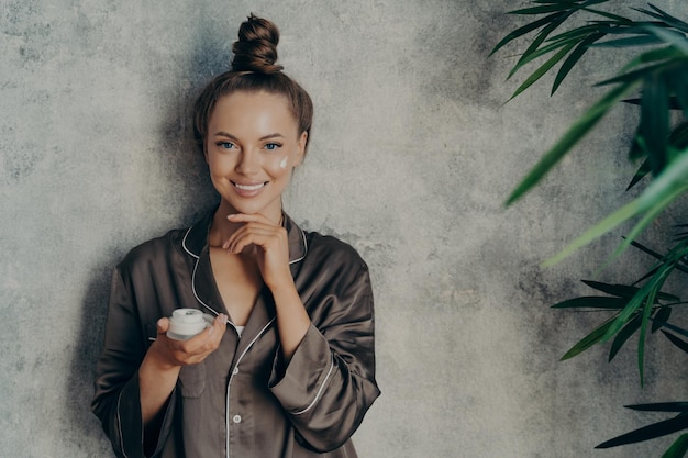 Una mujer feliz usa un pijama marrón de seda con crema hidratante en la mano durante la rutina de belleza matutina