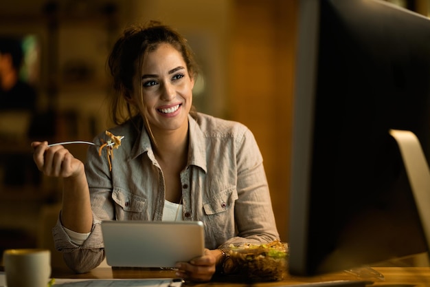 Mujer feliz usando tecnología inalámbrica mientras come ensalada por la noche en casa