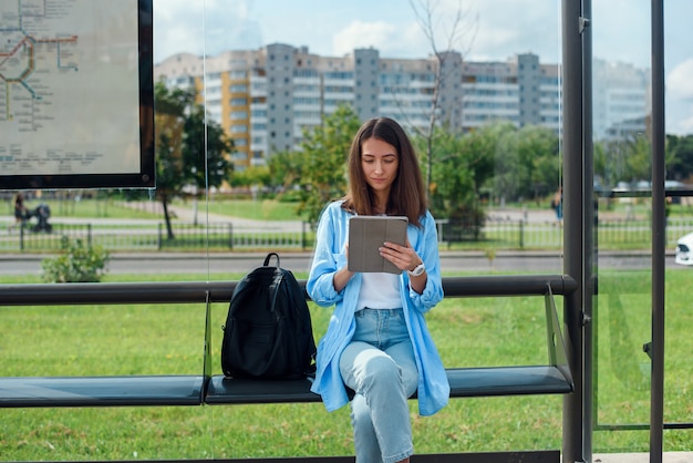 Mujer feliz utiliza una tableta o ebook en una estación de tranvía mientras espera el transporte público.