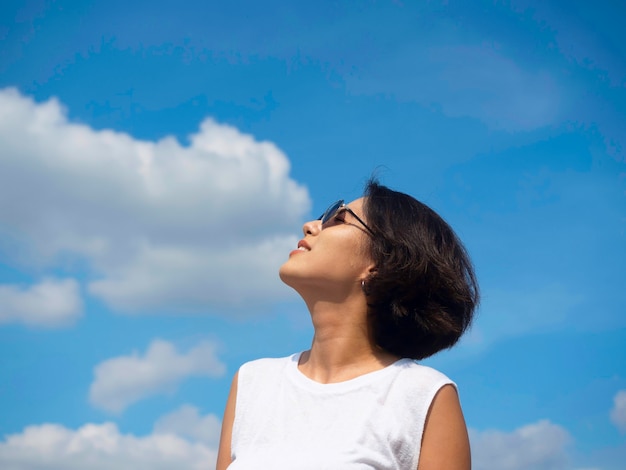 Mujer feliz en verano. Sonriente hermosa mujer asiática de pelo corto con gafas de sol y camisa blanca sin mangas casual mirando hacia el cielo azul y el fondo de nubes en un día soleado de verano.