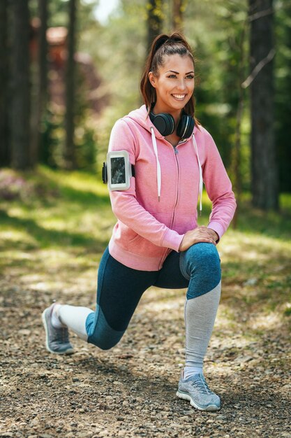 Mujer feliz, vestida con ropa deportiva, con auriculares alrededor del cuello, haciendo ejercicios de estocadas en un soleado sendero forestal, disfrutando del paisaje entre los árboles.