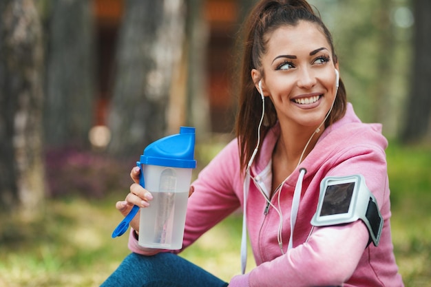 Mujer feliz, vestida con ropa deportiva, con auriculares en los oídos, está bebiendo agua en un descanso de ejercicios en un bosque soleado, disfrutando del paisaje entre los árboles.