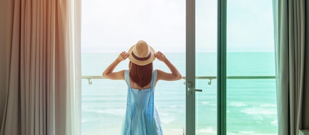 Mujer feliz con vestido azul y sombrero mirando por la ventana exterior a la vista del océano en la mañana Relájese en el tiempo de vacaciones para viajar y el concepto de libertad