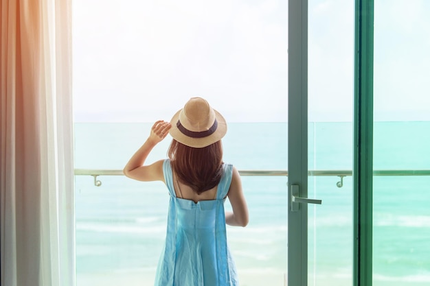 Mujer feliz con vestido azul y sombrero mirando por la ventana exterior a la vista del océano en la mañana Relájese en el tiempo de vacaciones para viajar y el concepto de libertad