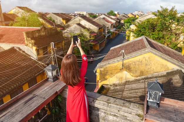 Mujer feliz con un vestido vietnamita Ao Dai viajero tomando fotos por teléfono móvil en la azotea en la antigua ciudad de Hoi An en Vietnam Vietnam y el concepto de viaje del sudeste
