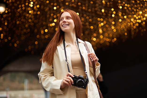 Mujer feliz viajera se encuentra al aire libre en un lugar decorado con luces, usa cámara retro, camina y toma fotos, luciendo feliz. Retrato de mujer caucásica pelirroja en chaqueta beige