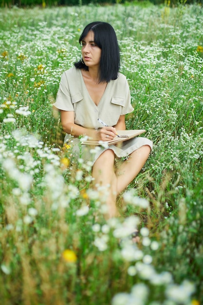 Mujer femenina con pluma escribiendo o pintando escritura a mano en un cuaderno en un prado floreciente