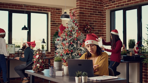 Mujer festiva hablando en una conferencia de videollamada en línea con colegas, asistiendo a una reunión de teleconferencia por Internet en una oficina decorada con árboles de Navidad y luces. Videoconferencia remota.