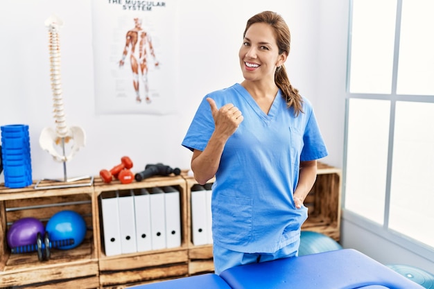 Mujer fisioterapeuta hispana de mediana edad que trabaja en la clínica de recuperación del dolor haciendo gestos felices con los pulgares hacia arriba con la mano. expresión de aprobación mirando a la cámara que muestra el éxito.