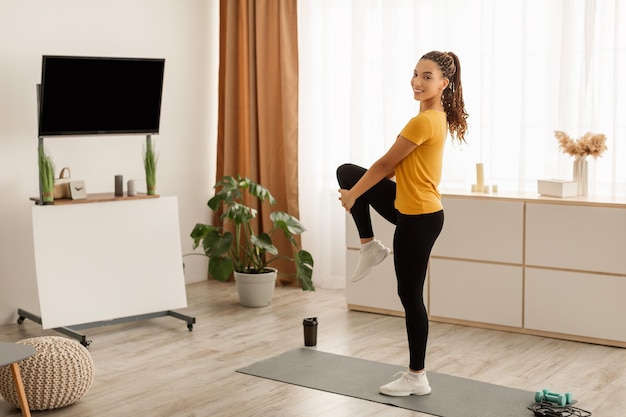 Mujer fitness afroamericana estirando la pierna haciendo ejercicio de pie en casa