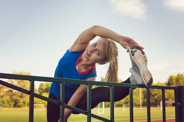 Mujer de fitness deporte que se extiende en el estadio. Deporte chica rubia estirando los brazos en la arena deportiva con muchas pistas. Ejercicio de la mañana de verano.