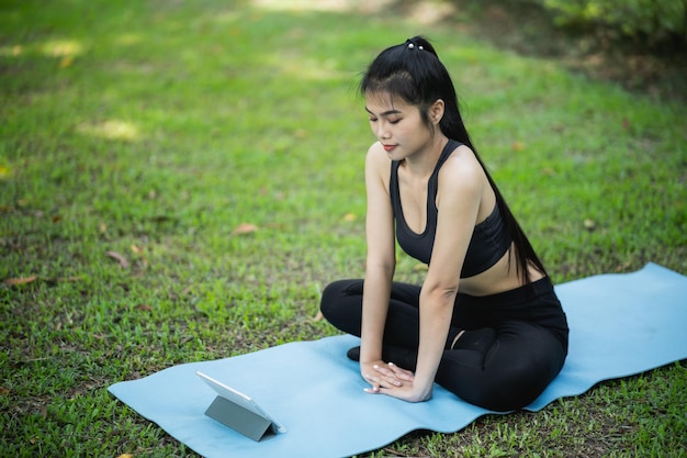 Mujer de fitness deportivo asiático aprendiendo haciendo yoga de acuerdo con un videoclip o videoconferencia con el maestro Mujer asiática usando tableta y haciendo yoga en el parque Estudio de yoga concepto de aprendizaje en línea