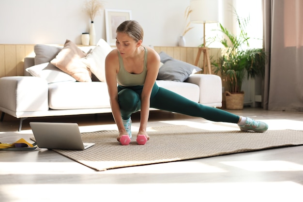 Mujer fitness haciendo ejercicios deportivos y viendo tutoriales en línea en la computadora portátil, entrenando en la sala de estar. Quédese en las actividades de la casa.