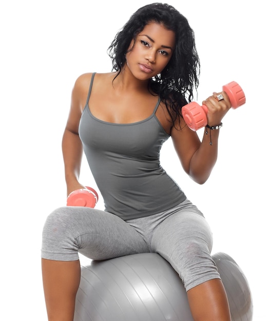 Mujer fitness morena con ropa deportiva gris sentada en una pelota de fitness y sosteniendo una mancuerna.