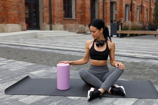 Mujer fitness en ropa deportiva sentada en una alfombra de yoga y toma un rodillo de masaje