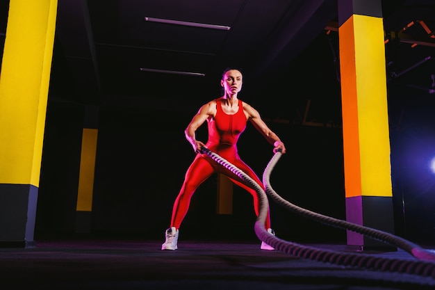 Mujer fitness utilizando cuerdas de entrenamiento para hacer ejercicio en el gimnasio. Atleta trabajando con cuerdas de batalla en el gimnasio de cross