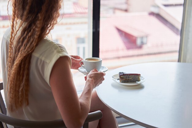 Mujer flaca pelirroja rizada sentada en la cafetería frente a la ventana