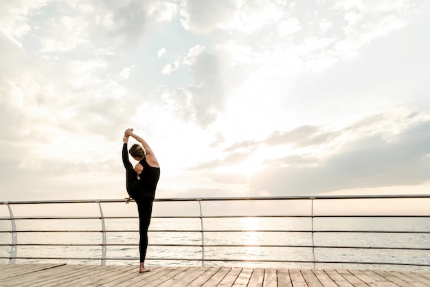 Mujer flexible haciendo asanas de yoga cerca del mar al amanecer de la mañana, practicando ejercicios deportivos y de fitness