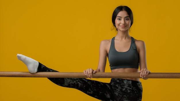 Mujer flexible con sonrisa étnica india disfruta estirándose en el estudio amarillo Bailarina acróbata dama deportiva chica sonriendo mirando a la cámara estira la pierna con barra de ballet ejercicio de flexibilidad