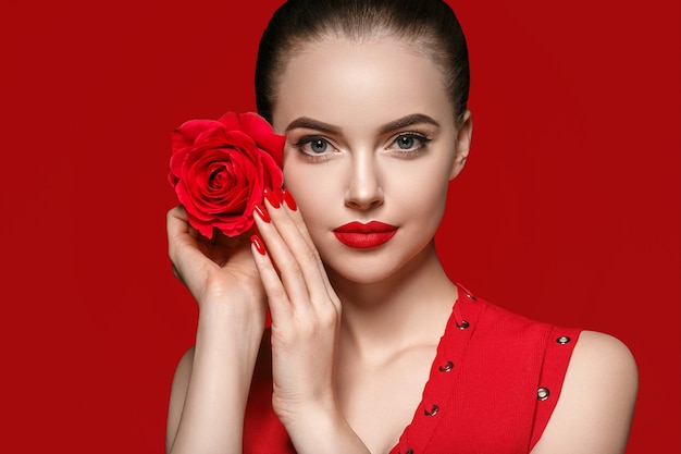 Mujer con flor color de rosa. Retrato de mujer de belleza con hermosa flor color de rosa y peinado de salón sobre fondo rojo. Tiro del estudio.