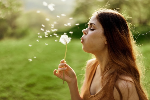 Mujer con una flor de diente de león en sus manos sonriendo y soplando sobre un fondo de vegetación de verano y luz solar