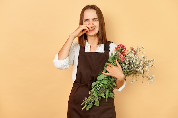 Mujer florista enferma con un delantal marrón sosteniendo un ramo de flores de pie aislada sobre un fondo beige sufriendo alergia estacional mientras trabaja en una floristería