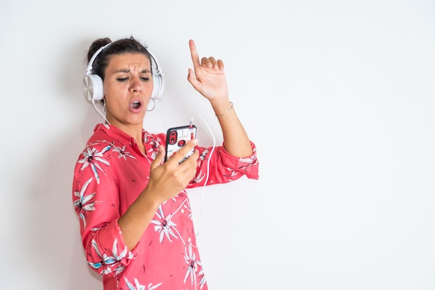 Una mujer de fondo blanco apunta a su teléfono y lleva auriculares cantando