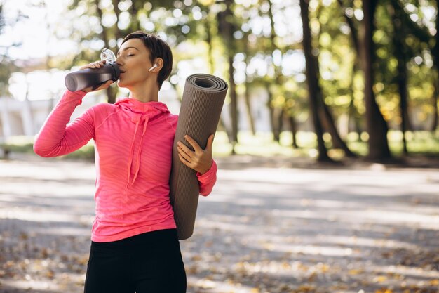 Mujer en forma bebiendo agua en el parque y sosteniendo estera de yoga