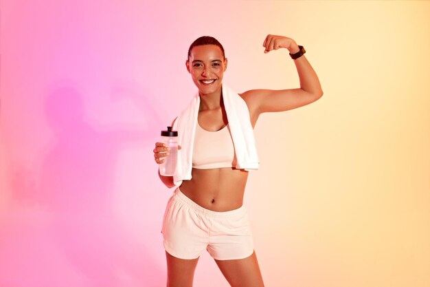 Una mujer en forma con la cabeza afeitada flexiona con orgullo su bíceps sosteniendo una botella de agua y una toalla