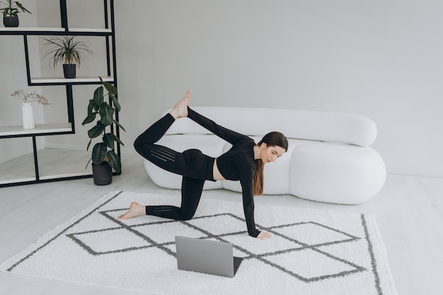 Mujer en forma haciendo yoga y viendo tutoriales en línea sobre entrenamiento de portátiles en la sala de estar