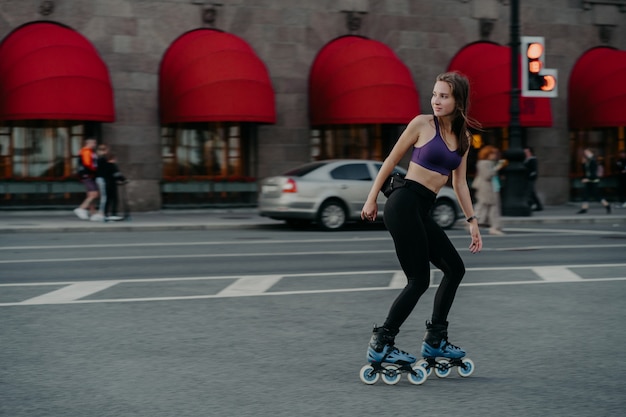 Mujer en forma involucrada en patinaje sobre ruedas usa top y poses de leggings en una carretera concurrida involucrada en deporte recreativo popular mira hacia otro lado con expresión pensativa intenta perder peso aumenta la salud emocional